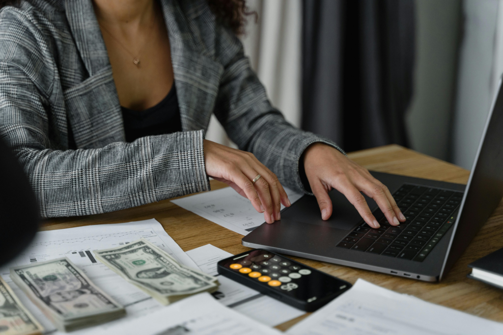 A Finance Professional is seated at a desk, focused on her laptop computer. Her hands are on the keyboard, typing away. Beside her, there is a stack of money neatly placed on the table. The room is well lit, allowing her to work comfortably. In the background, a remote control can be seen resting on the desk. The woman is wearing a ring on her finger and a necklace around her neck. The scene conveys a sense of productivity and focus as she attends to her work tasks.