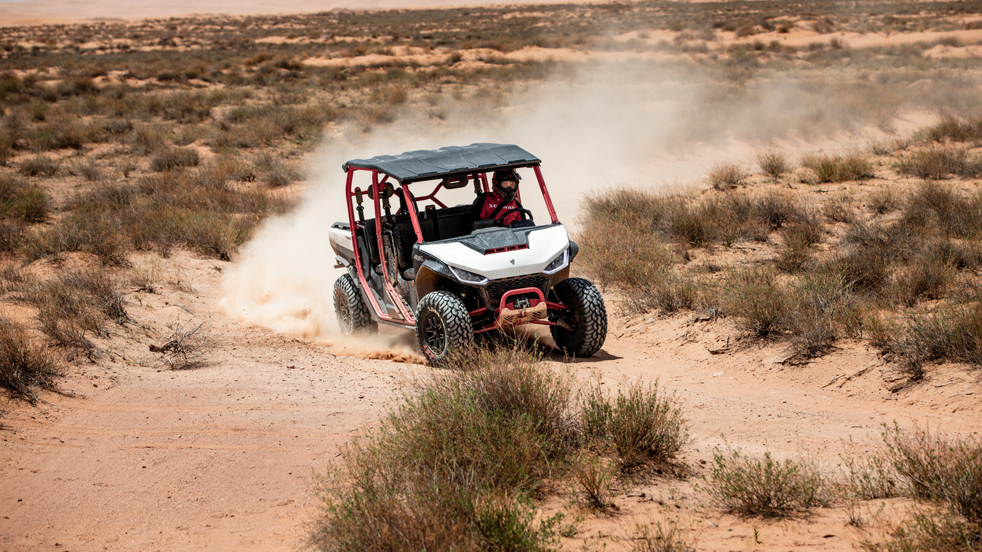 A man is seen driving a Segway UTV (utility task vehicle) through a vast desert landscape. The UTV is a rugged SUV-like vehicle designed for off-road use, and it is kicking up dust as it travels along a sandy road. The man is wearing a red jacket and a helmet, indicating that he is taking safety precautions while driving. The scene is characterized by the arid desert environment, with dunes and sand stretching out in all directions. The image captures the adventurous spirit of exploring remote desert terrains in a sturdy off-road vehicle.